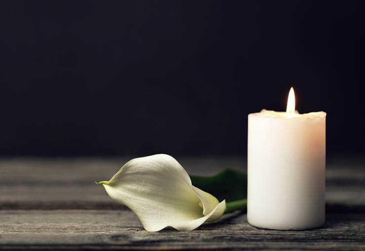 A somber image depicts a lit candle and a white calla lily resting on a wooden surface.